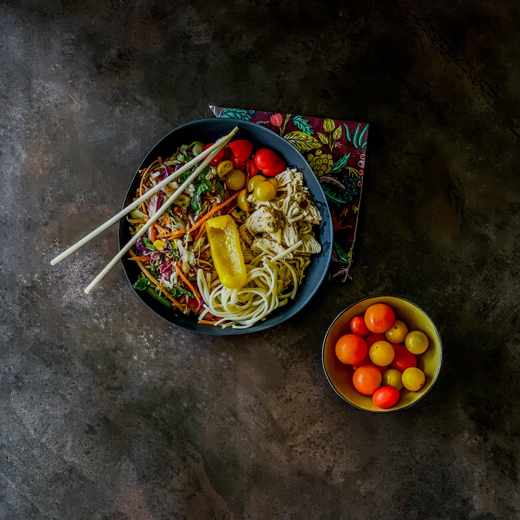 Colourful bowl of noodles with vegetables and a side of cherry tomatoes on a dark photography backdrop from Flatlay Studio.